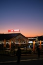 A cozy cafe with a string of warm lights creating a welcoming ambiance at dusk. The sky is a gradient of blue and orange, indicating sunset. The cafe has a wooden structure with large windows, and several umbrellas are closed on the patio. A prominent sign with the words 'Cafe Tourist' is illuminated on the roof.