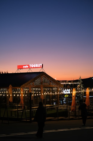 A cozy cafe with a string of warm lights creating a welcoming ambiance at dusk. The sky is a gradient of blue and orange, indicating sunset. The cafe has a wooden structure with large windows, and several umbrellas are closed on the patio. A prominent sign with the words 'Cafe Tourist' is illuminated on the roof.