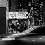 A black and white photograph depicts a bustling urban scene, featuring a theater marquee with a crowd gathered around the entrance. The marquee advertises a show titled 'Who's Live Anyway' scheduled for 730 PM. The street is busy with blurred car lights indicating motion, and tall buildings are visible in the background, illuminated against the night sky.