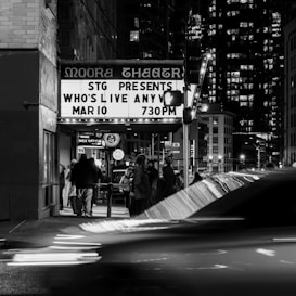 A black and white photograph depicts a bustling urban scene, featuring a theater marquee with a crowd gathered around the entrance. The marquee advertises a show titled 'Who's Live Anyway' scheduled for 730 PM. The street is busy with blurred car lights indicating motion, and tall buildings are visible in the background, illuminated against the night sky.