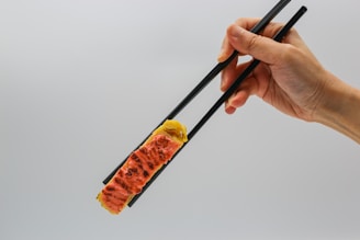 A hand is holding black chopsticks, gripping a piece of sushi that is topped with a slightly grilled slice of fish. The background is a plain light color, providing a minimalistic setting.