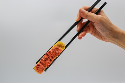 A hand is holding black chopsticks, gripping a piece of sushi that is topped with a slightly grilled slice of fish. The background is a plain light color, providing a minimalistic setting.