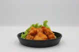 A black plate holds a serving of crispy, golden-brown fried chicken. The chicken pieces are arranged nicely, resting on a bed of fresh green lettuce leaves, which add a touch of color and freshness to the dish. The background is a neutral, soft white, highlighting the vibrant colors of the food.