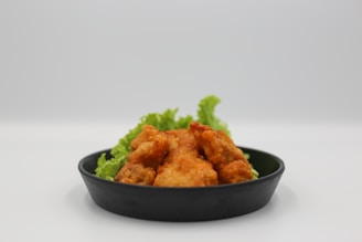 A black plate holds a serving of crispy, golden-brown fried chicken. The chicken pieces are arranged nicely, resting on a bed of fresh green lettuce leaves, which add a touch of color and freshness to the dish. The background is a neutral, soft white, highlighting the vibrant colors of the food.
