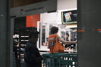 Inside a store, a person wearing a basketball jersey with the number 23 is standing at the counter. Another person behind the counter appears to be assisting them. The setting includes shelves with products and a lit sign advertising a snack item.