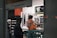 Inside a store, a person wearing a basketball jersey with the number 23 is standing at the counter. Another person behind the counter appears to be assisting them. The setting includes shelves with products and a lit sign advertising a snack item.