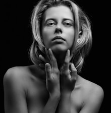A black and white portrait of a person with shoulder-length hair, looking directly at the camera. The individual holds their hands gently around their neck, creating a composed and contemplative pose. The lighting accentuates facial features and creates shadows, adding depth to the image.
