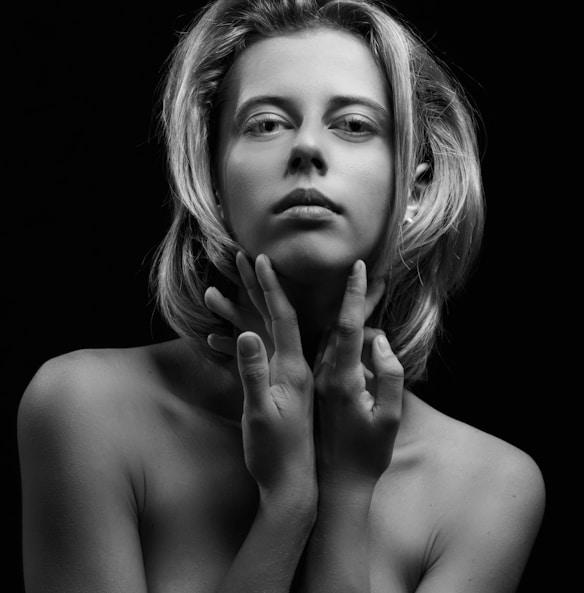 A black and white portrait of a person with shoulder-length hair, looking directly at the camera. The individual holds their hands gently around their neck, creating a composed and contemplative pose. The lighting accentuates facial features and creates shadows, adding depth to the image.