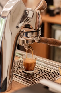 A coffee machine is dispensing an espresso shot into a small glass. The machine has a metallic and wooden handle design, and the espresso is flowing into a clear glass on a drip tray.