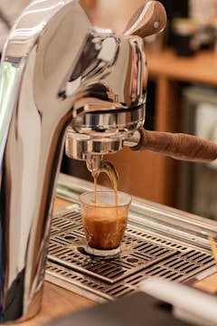A coffee machine is dispensing an espresso shot into a small glass. The machine has a metallic and wooden handle design, and the espresso is flowing into a clear glass on a drip tray.