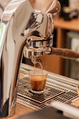 A coffee machine is dispensing an espresso shot into a small glass. The machine has a metallic and wooden handle design, and the espresso is flowing into a clear glass on a drip tray.