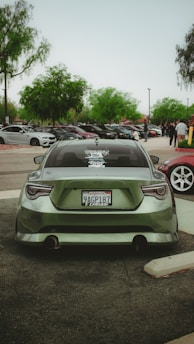 A green sports car is parked in a parking lot with several other vehicles visible in the background. The car has a distinct rear design and a modified body. Trees with lush green foliage are scattered around the parking lot, and a group of people is gathered in the vicinity.