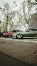 A low-angle view of parked cars, with a focus on two modified, sporty vehicles. One car is red with a sleek design, and the other is green, featuring low-profile tires and custom rims. The background shows tall trees with sparse leaves, and part of a modern building.
