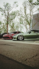 A low-angle view of parked cars, with a focus on two modified, sporty vehicles. One car is red with a sleek design, and the other is green, featuring low-profile tires and custom rims. The background shows tall trees with sparse leaves, and part of a modern building.