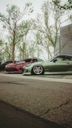 A low-angle view of parked cars, with a focus on two modified, sporty vehicles. One car is red with a sleek design, and the other is green, featuring low-profile tires and custom rims. The background shows tall trees with sparse leaves, and part of a modern building.