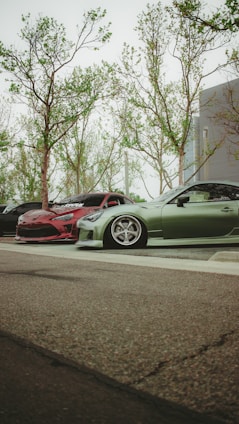A low-angle view of parked cars, with a focus on two modified, sporty vehicles. One car is red with a sleek design, and the other is green, featuring low-profile tires and custom rims. The background shows tall trees with sparse leaves, and part of a modern building.