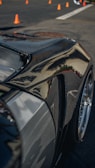 A close-up of a sleek, polished car body, likely made of carbon fiber or with a glossy finish, reflecting light and surroundings. Orange traffic cones are visible in the background on a paved surface, suggesting a controlled environment like a driving course or a car show.