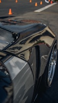 A close-up of a sleek, polished car body, likely made of carbon fiber or with a glossy finish, reflecting light and surroundings. Orange traffic cones are visible in the background on a paved surface, suggesting a controlled environment like a driving course or a car show.