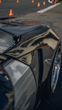 A close-up of a sleek, polished car body, likely made of carbon fiber or with a glossy finish, reflecting light and surroundings. Orange traffic cones are visible in the background on a paved surface, suggesting a controlled environment like a driving course or a car show.