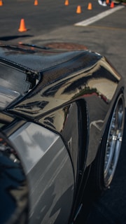 A close-up of a sleek, polished car body, likely made of carbon fiber or with a glossy finish, reflecting light and surroundings. Orange traffic cones are visible in the background on a paved surface, suggesting a controlled environment like a driving course or a car show.