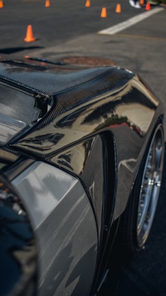 A close-up of a sleek, polished car body, likely made of carbon fiber or with a glossy finish, reflecting light and surroundings. Orange traffic cones are visible in the background on a paved surface, suggesting a controlled environment like a driving course or a car show.