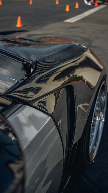 A close-up of a sleek, polished car body, likely made of carbon fiber or with a glossy finish, reflecting light and surroundings. Orange traffic cones are visible in the background on a paved surface, suggesting a controlled environment like a driving course or a car show.