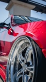 A close-up view of a polished chrome car wheel with reflective surfaces, set against a shiny red vehicle body. The image highlights the sleek design and glossy finish, with sunlight reflecting off the metallic surfaces.