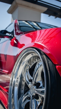 A close-up view of a polished chrome car wheel with reflective surfaces, set against a shiny red vehicle body. The image highlights the sleek design and glossy finish, with sunlight reflecting off the metallic surfaces.