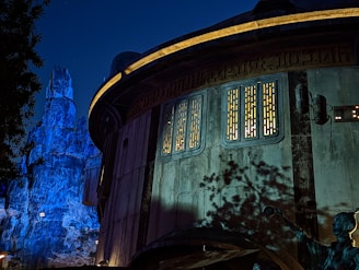 A futuristic, round building with illuminated panels and intricate designs, set against a nighttime backdrop. The structure features sci-fi elements such as exposed cables and a control panel. The background includes rocky, mountainous terrain bathed in blue light, enhancing the otherworldly ambiance. Shadows of nearby foliage are visible on the building's surface.