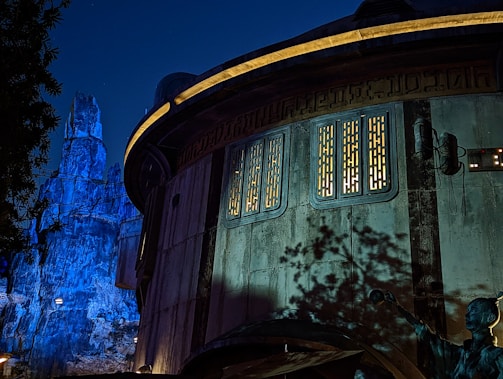 A futuristic, round building with illuminated panels and intricate designs, set against a nighttime backdrop. The structure features sci-fi elements such as exposed cables and a control panel. The background includes rocky, mountainous terrain bathed in blue light, enhancing the otherworldly ambiance. Shadows of nearby foliage are visible on the building's surface.