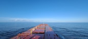 A large cargo ship sails through calm, open waters under a clear blue sky. The expansive deck stretches forward, showing a helipad and various containers secured on board.