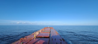 A large cargo ship sails through calm, open waters under a clear blue sky. The expansive deck stretches forward, showing a helipad and various containers secured on board.
