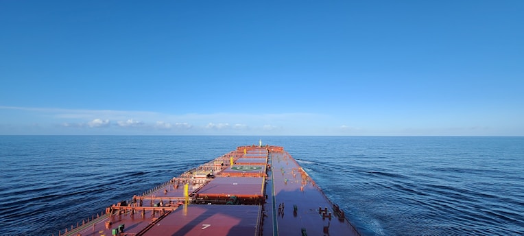 A large cargo ship sails through calm, open waters under a clear blue sky. The expansive deck stretches forward, showing a helipad and various containers secured on board.
