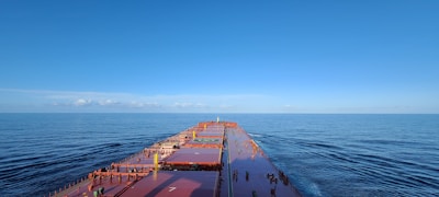 A large cargo ship sails through calm, open waters under a clear blue sky. The expansive deck stretches forward, showing a helipad and various containers secured on board.