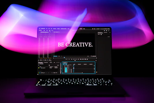 A laptop is displayed with a video editing software open on the screen. The words 'Be Creative.' appear prominently, suggesting a focus on creativity. The background features swirling light patterns in shades of purple, pink, and blue, creating an artistic ambiance.