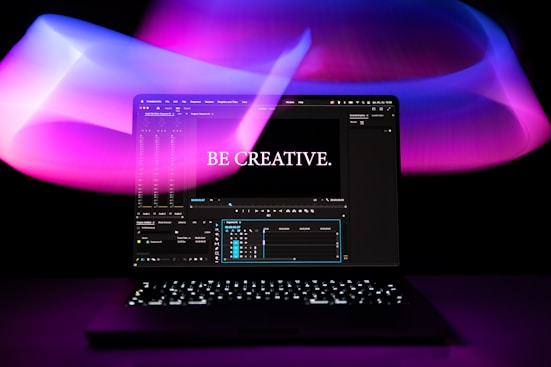 A laptop is displayed with a video editing software open on the screen. The words 'Be Creative.' appear prominently, suggesting a focus on creativity. The background features swirling light patterns in shades of purple, pink, and blue, creating an artistic ambiance.