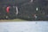 A group of kite surfers is gliding across a wide, blue lake surrounded by dense green forests. Several colorful kites are in the air with a waterfall visible in the background.