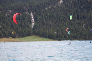 A group of kite surfers is gliding across a wide, blue lake surrounded by dense green forests. Several colorful kites are in the air with a waterfall visible in the background.