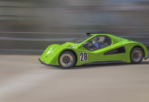 A bright green race car with the number 28 on its side is speeding on a track. The background is blurred, emphasizing the car's motion. The driver is looking forward and wearing a helmet.