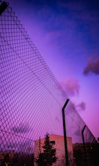 A tall chain-link fence against a vibrant purple and blue sky with scattered clouds. In the background, buildings and trees can be seen, partially obscured by the fence.