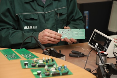 A person wearing a dark green work jacket is holding a green printed circuit board. The person appears to be at a workstation with electronic components and tools. There are several green circuit boards on the wooden table, along with a soldering iron, wire cutters, and a spool of soldering wire. An electronic device with a display is also present on the desk.