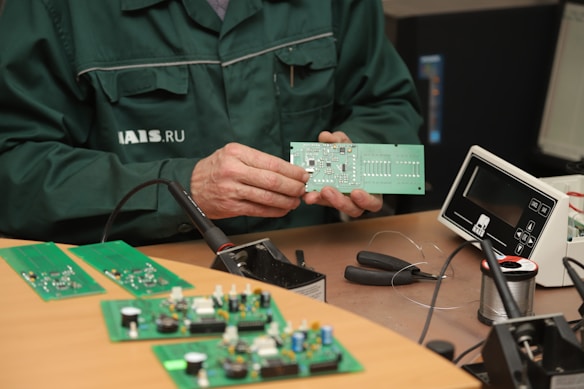 A person wearing a dark green work jacket is holding a green printed circuit board. The person appears to be at a workstation with electronic components and tools. There are several green circuit boards on the wooden table, along with a soldering iron, wire cutters, and a spool of soldering wire. An electronic device with a display is also present on the desk.