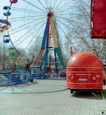 A vibrant amusement park scene featuring a large Ferris wheel with colorful gondolas and a bright red food truck shaped like a trailer. The Ferris wheel has multiple colors on its supports consisting of blue, red, green, and yellow. Bare trees can be seen in the background, suggesting an off-season or early spring setting. The ground is paved with light stone tiles, and there are various fences and entry gates visible.