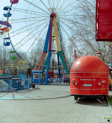 A vibrant amusement park scene featuring a large Ferris wheel with colorful gondolas and a bright red food truck shaped like a trailer. The Ferris wheel has multiple colors on its supports consisting of blue, red, green, and yellow. Bare trees can be seen in the background, suggesting an off-season or early spring setting. The ground is paved with light stone tiles, and there are various fences and entry gates visible.