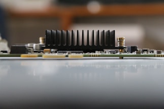 A close-up side view of an electronic circuit board featuring a prominent black heat sink with vertical fins. The board contains small electronic components, including resistors and capacitors, with a blurred background.