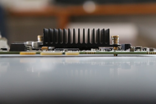 A close-up side view of an electronic circuit board featuring a prominent black heat sink with vertical fins. The board contains small electronic components, including resistors and capacitors, with a blurred background.