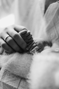 A black and white close-up of an adult hand gently holding a baby's tiny feet. The adult is wearing a ring and the baby is wrapped in a soft, fuzzy fabric.