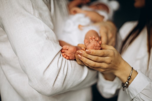 A close-up view of a baby's feet being gently held by a pair of adult hands. The baby is wearing a soft white outfit, and the adults are dressed in light-colored clothing. The scene conveys a sense of tenderness and care.