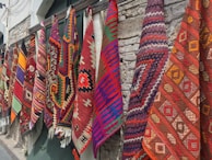 Several colorful and intricately patterned rugs hang side by side against a stone wall. The patterns include geometric shapes and are woven in vibrant colors such as red, orange, purple, blue, and green.