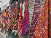 Several colorful and intricately patterned rugs hang side by side against a stone wall. The patterns include geometric shapes and are woven in vibrant colors such as red, orange, purple, blue, and green.